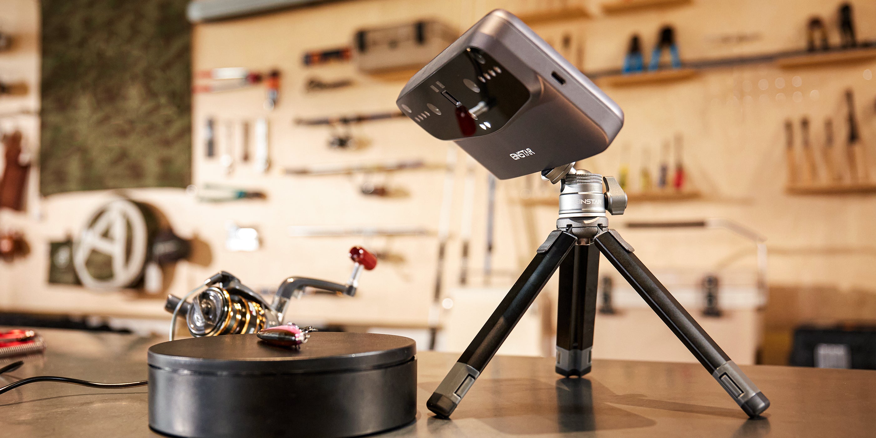 EINSTAR VEGA 3D scanner mounted on a tripod, positioned next to a rotating platform for object scanning in a workshop setting. The durable metal body of the tripod and scanner highlights the sleek and refined design, emphasizing build quality.